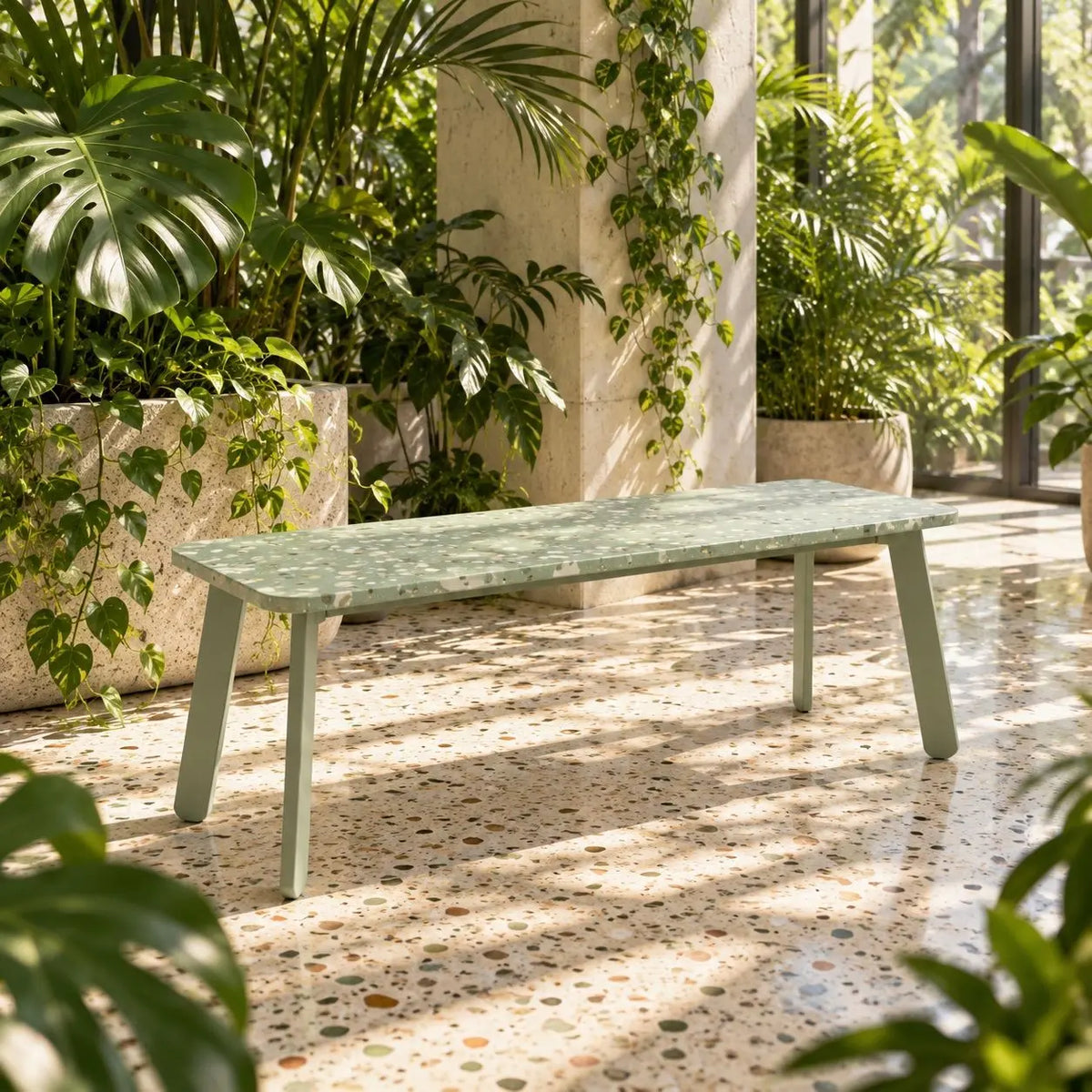 A long rectangular bench with a speckled sage green stone top and matching legs, placed in a bright indoor garden setting with potted plants and terrazzo flooring. Bench made from recycled plastic panels.