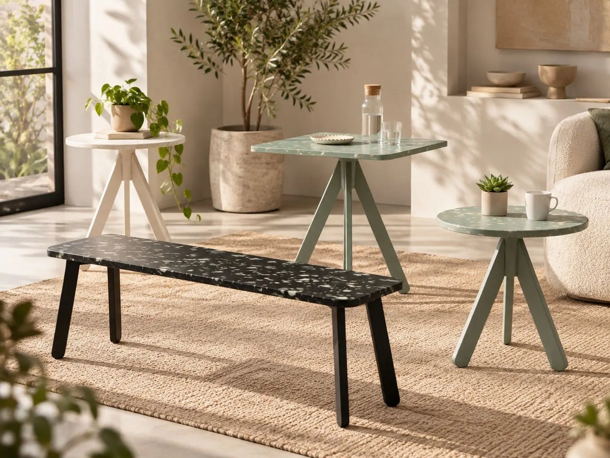 A styled interior scene showing a black marble-top bench with dark legs, a mint green square table, and a mint green round side table in a bright living room with plants and neutral decor. Furniture made from recycled plastic panels.