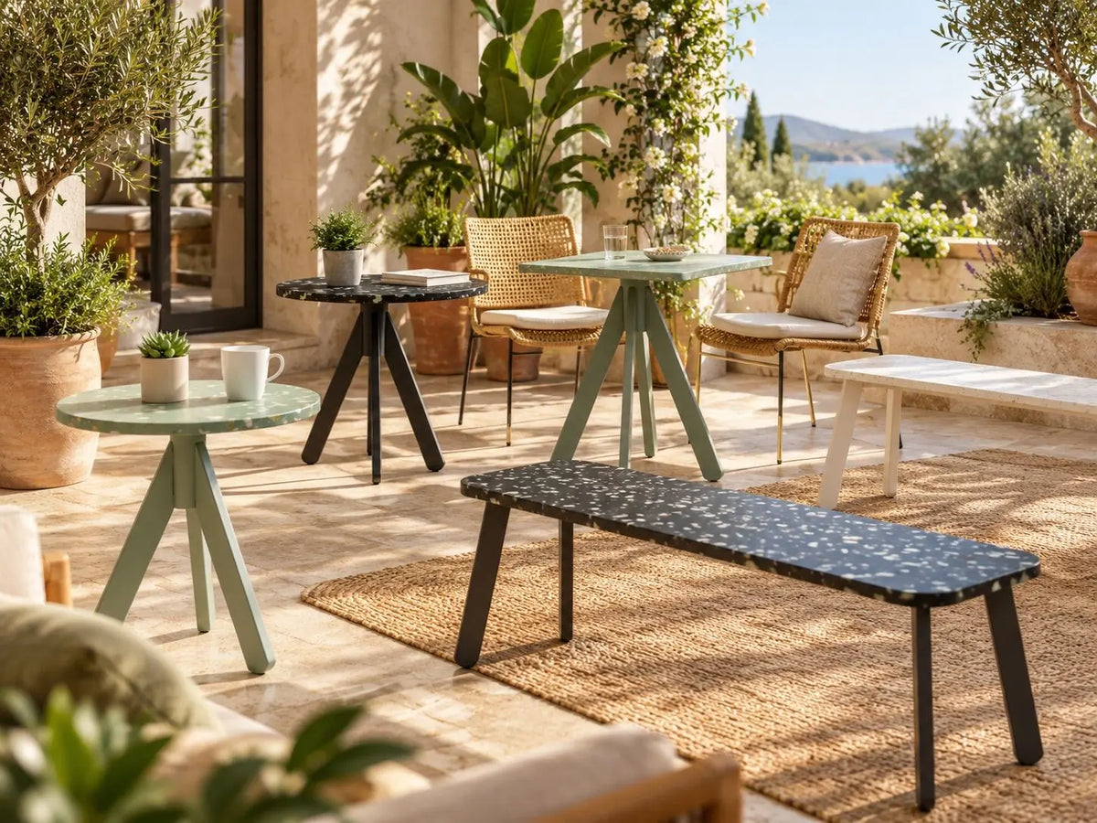 Outdoor patio furniture set including a long bench, round tables, and woven chairs on a stone terrace with plants and a scenic background. Furinutre made from recycled plastic panels.