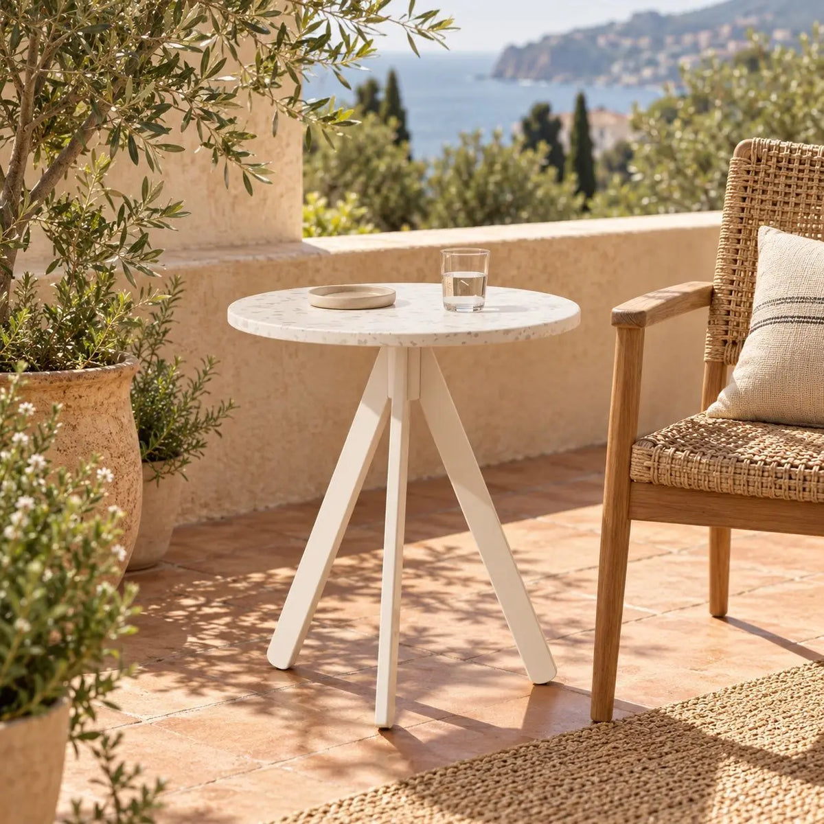 A small round outdoor side table with a white stone-look top and white tripod base, styled on a terracotta patio beside a woven chair with a glass and small dish on top. The setting includes potted plants, a stucco wall, and a scenic coastal view in the background. Table made from recycled plastic panels.
