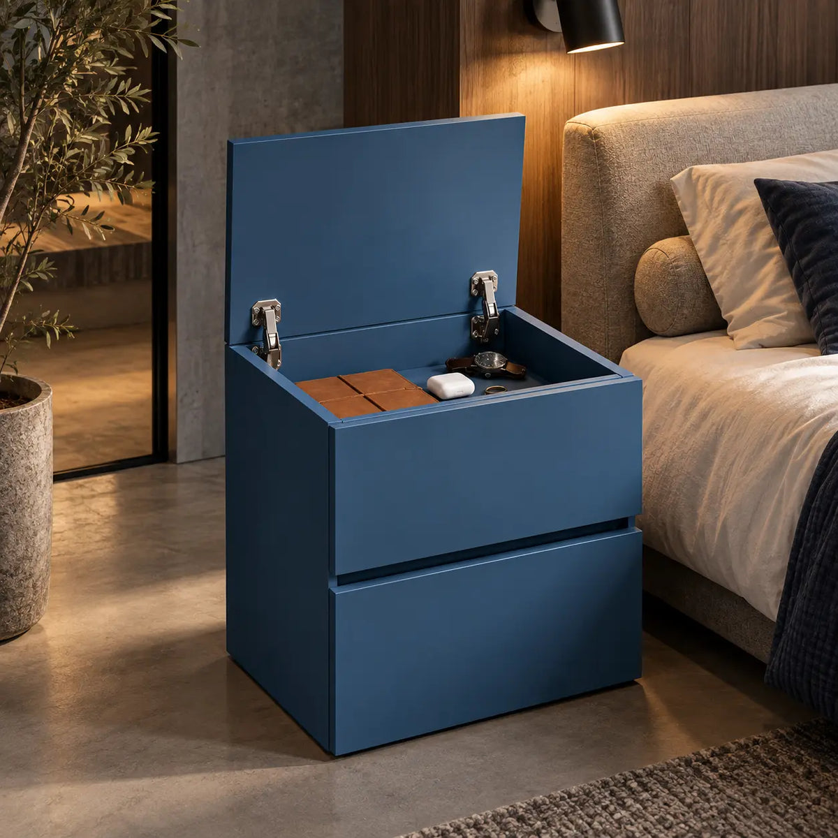 A blue storage bedside table with a lift-up lid, shown in a modern bedroom setting next to a bed with beige bedding. Concealted storage furniture.