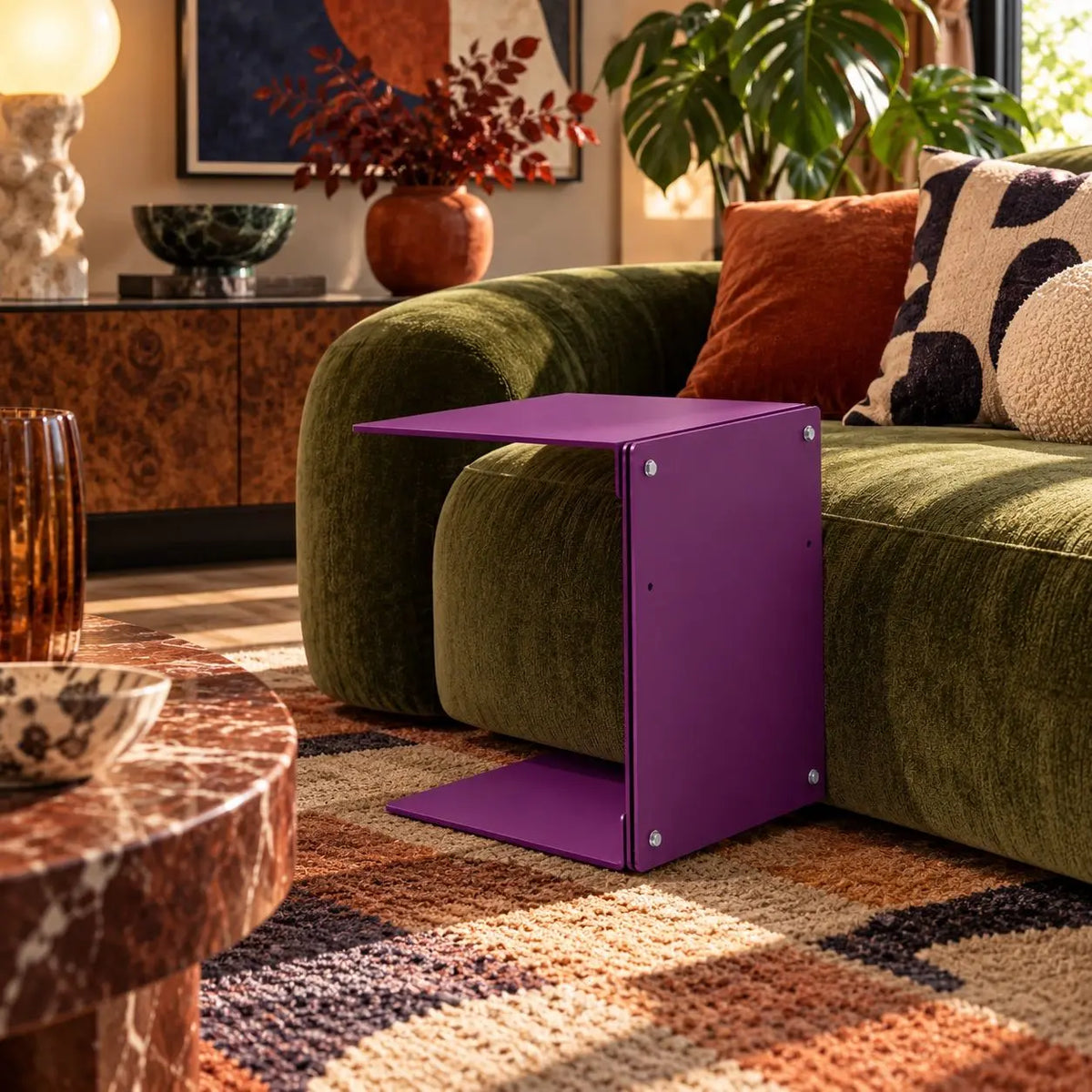A modern purple side table with a geometric, folded-panel design stands next to a green velvet sofa in a styled living room. Aluminum high quality side table. 