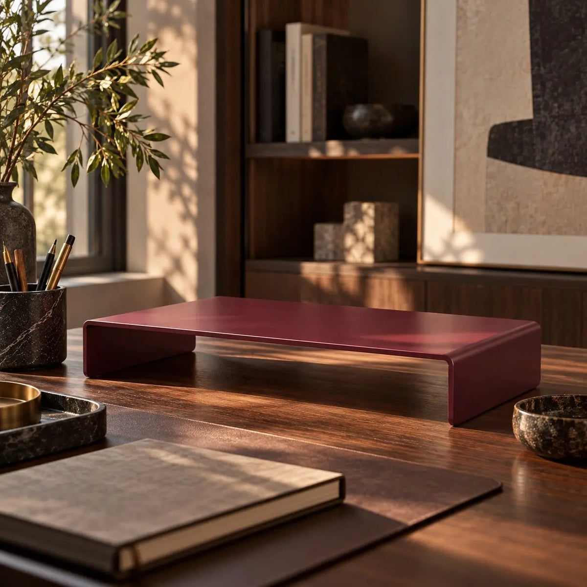 A modern, minimalist rectangular desk monitor stand with a matte burgundy-red top and silver metal legs, styled on a wooden desk in a warm, sunlit room. Aluminum high quality monitor stand. 