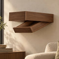 A floating wall-mounted wooden shelf with a lower pull-out tray, shown installed on a beige wall in a styled interior with part of a side table, books, a plant, and a chair visible. Hidden compartment.