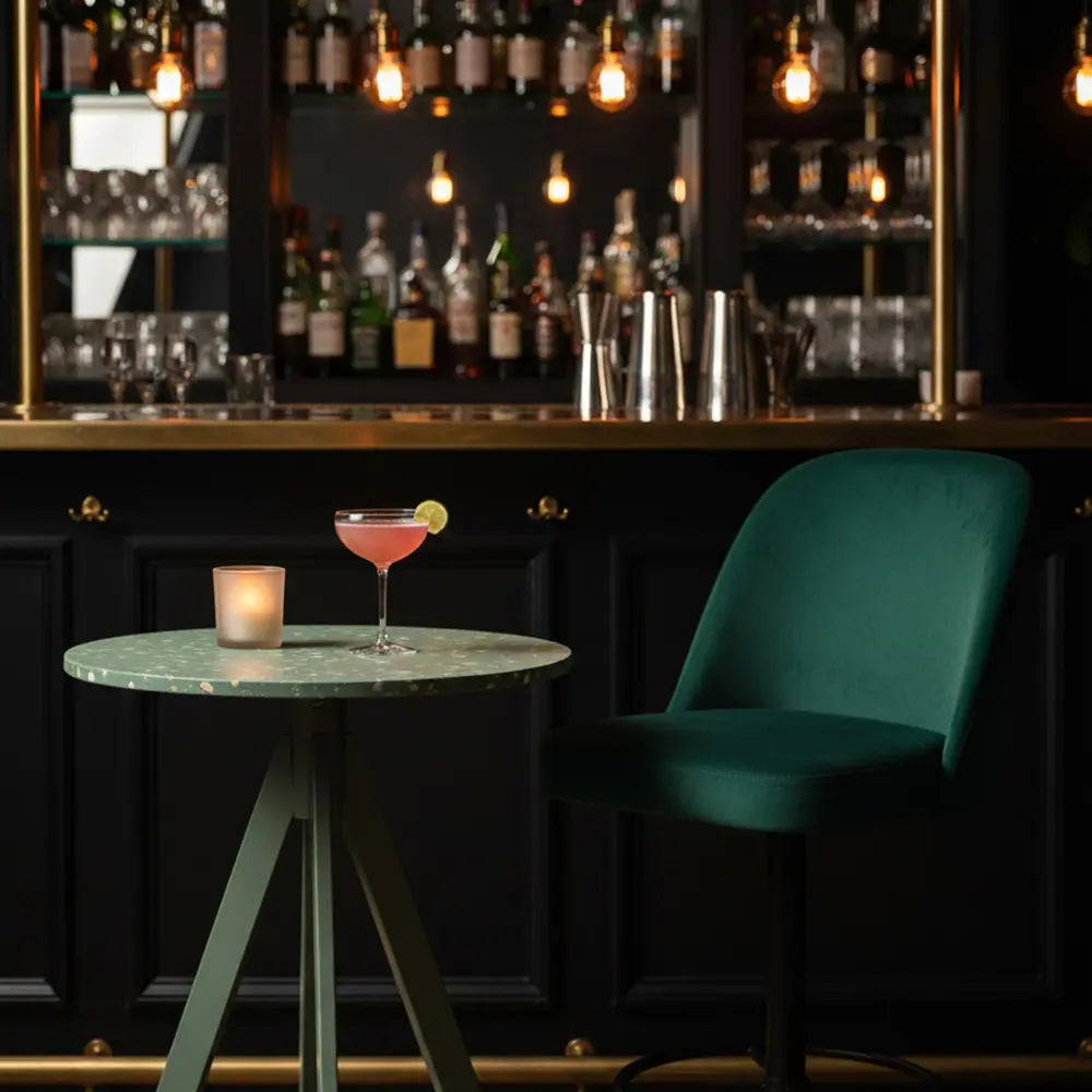 Round green recycled HDPE table in a cocktail bar, paired with a green velvet chair and a garnished pink cocktail