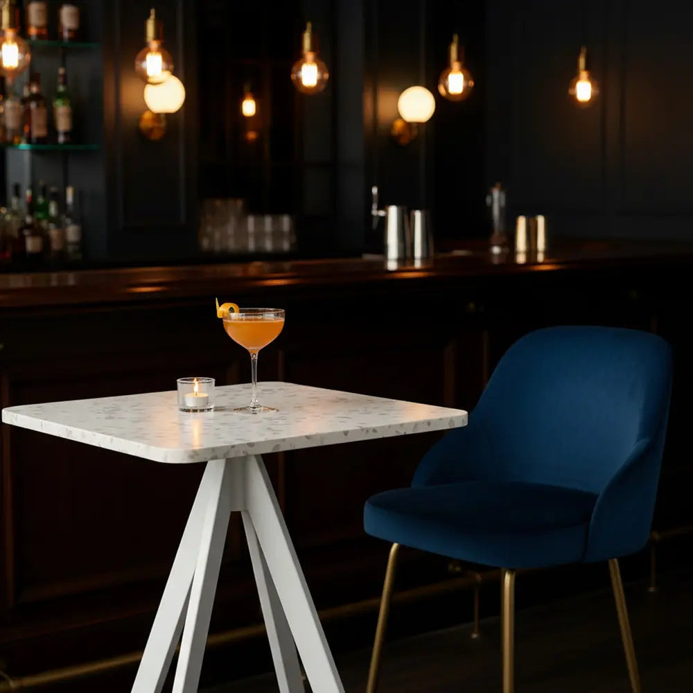 Square white eco-friendly table made from recycled plastic in a dark cocktail bar, styled with a blue velvet chair, candle, and garnished drink